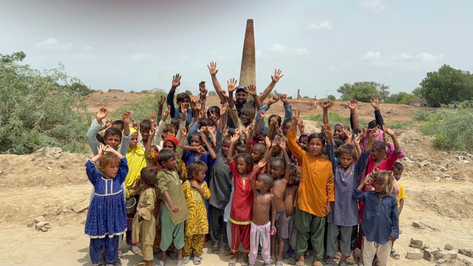 A huge crowd of children at a brick kiln raise their hands and cheer
