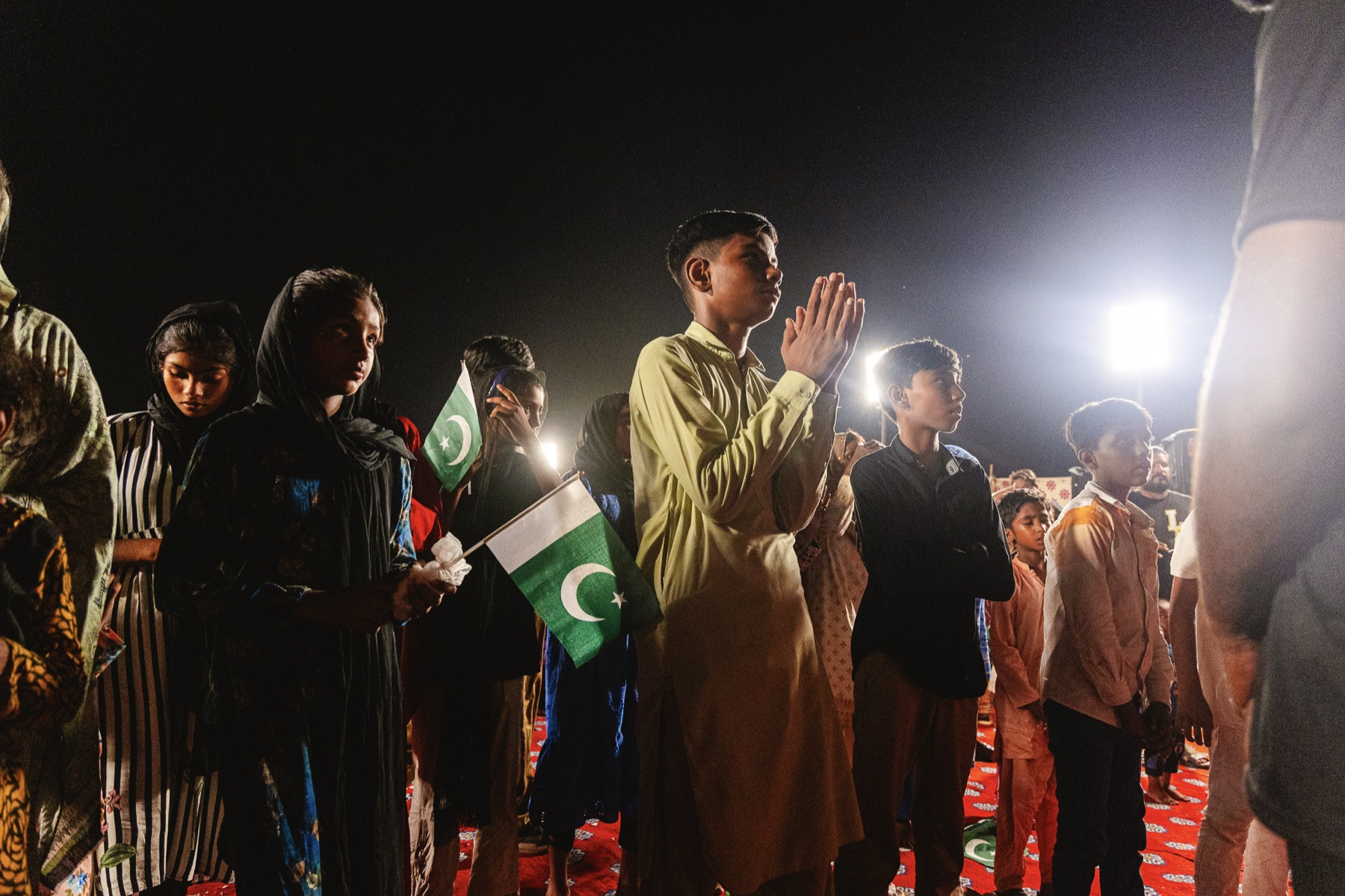 Families pray at a night gathering, holding Pakistani flags