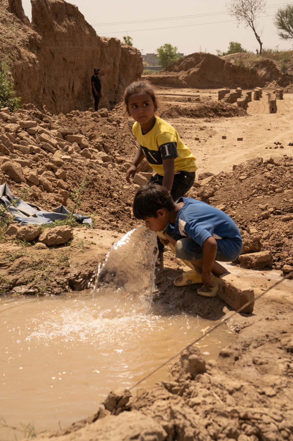 Children drink from a muddy water source beside a brick kiln